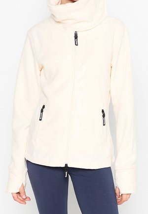 Vrouw draagt een crèmekleurig fleecevest met rits en hoge kraag, gecombineerd met marineblauwe leggings, handen ontspannen langs het lichaam.