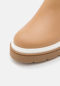 Avant de chaussure en cuir beige avec semelle épaisse marron et bordure blanche, montrant une texture lisse et un design à bout arrondi.