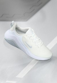 Hvid sportsko med et struktureret mesh-overdel, flade snørebånd og en lyseblå swoosh. Har en rund tå og en polstret sål.