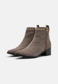 Botas de tobillo de ante gris con tachuelas doradas y cremalleras, con punta afilada y tacón bajo, exhibidas sobre un fondo blanco liso.