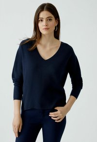 Pull en V bleu marine avec des manches longues, en tissu tricot texturé, légèrement tombé aux épaules, associé à un pantalon bleu foncé.