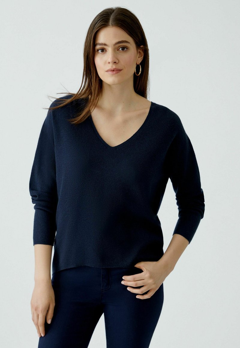 Pull en V bleu marine avec des manches longues, en tissu tricot texturé, légèrement tombé aux épaules, associé à un pantalon bleu foncé.