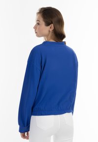 Fényes kék, hosszú ujjú pulóver, laza szabású, bordás mandzsettákkal és enyhén cropped fazonnal, puha anyagból készült.