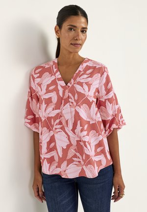 Vrouw met donker haar in een knot draagt een roze bloemen V-hals blouse met ellebooglange mouwen en donkerblauwe spijkerbroek, staand tegen een witte muur.