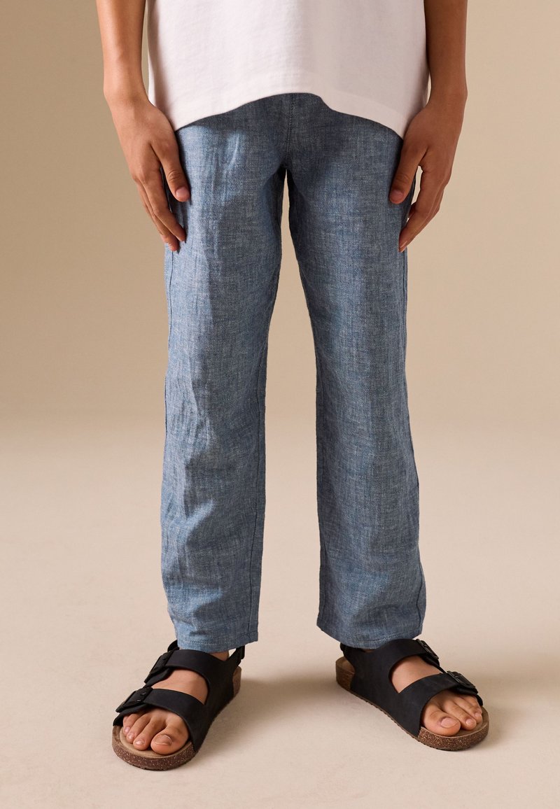 Niño con pantalones de lino azules, camisa blanca y sandalias negras de pie sobre un suelo beige frente a un fondo liso.