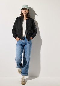 Svart bomberjacka, vit t-shirt, ljusblå vida jeans och beige sneakers med vita snörningar. Ljusgrön keps ovanför. Platt bakgrund.