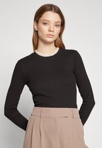 Svart langermet topp med ribbet tekstur og hvit volangkrage, kombinert med høy midje beige bukser, vist mot en nøytral bakgrunn.