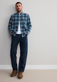 Cămașă flanel în carouri albastru și negru, tricou alb, pantaloni din denim închisi la culoare și cizme maro cu șireturi și talpă texturată. Îmbrăcăminte casual.