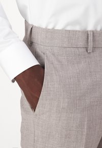 Calças de linho cinza claro com uma textura suave, apresentando um bolso lateral visível e uma cintura com passantes para cinto, acompanhadas de uma manga de camisa branca.