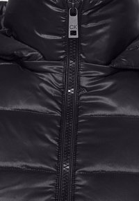 Černá péřová bunda s lesklým povrchem, obsahující centrální zip a prošívané panely. Kapucí design s pevnou šňůrkou na zip.