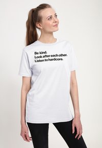 Bílý bavlněný tričko s odvážným černým textem: "Buďte laskaví. Pečujte o sebe navzájem. Poslouchejte hardcore." Unisex střih s krátkým rukávem.