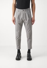Pantalons gris ajustés avec un léger éclat, dotés d'une taille élastique, de plis à l'avant et d'une longueur cheville. Portés avec des chaussures et des chaussettes noires.