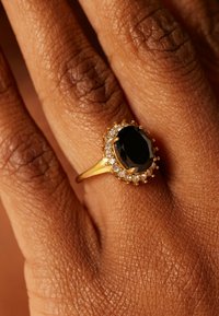 Gouden ring met een grote, ovale zwarte steen omringd door kleine heldere stenen, rustend op een getextureerde bruine hand.