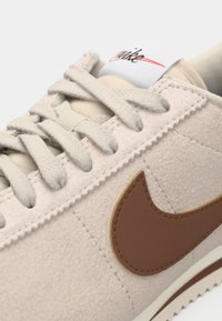 Gros plan sur une chaussure Nike beige avec un tissu texturé, un logo Swoosh marron et des lacets beiges assortis noués sur le dessus.
