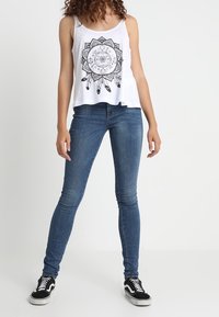Débardeur blanc avec impression mandala noire, associé à un jean skinny bleu foncé et des baskets noir et blanc. Design plat avec bretelles fines.