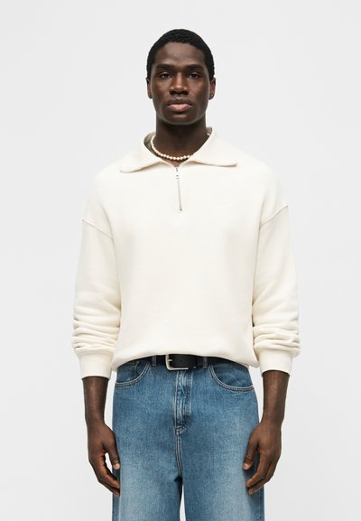 Άνδρας που φοράει ένα κρεμ πουλόβερ με φερμουάρ έως το μέσο, μπλε τζιν με μαύρη ζώνη και ένα κολιέ από μαργαριτάρια, στέκεται μπροστά σε απλό φόντο.