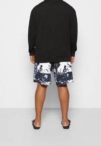 Sweatshirt noir à manches longues associé à un short de bain à motifs en blanc, noir et nuances de bleu. Des sandales avec une semelle noire sont portées.