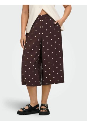 Femeie purtând pantaloni scurți evazați de culoare violet închis cu buline albe, un top alb fără mâneci și sandale negre cu catarame.