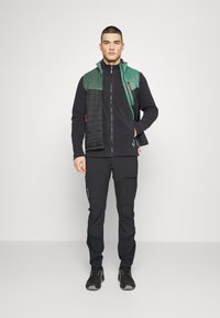 Schwarze Fleecejacke mit einer grünen, gesteppte Weste darüber. Dazu passende schwarze Hose und robustes schwarzes Schuhwerk. Mit Reißverschlüssen und Taschen ausgestattet.