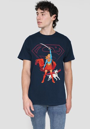Young man wearing navy blue T-shirt featuring Supergirl holding a sword with a white dog at her feet, standing in front of a large emblem.