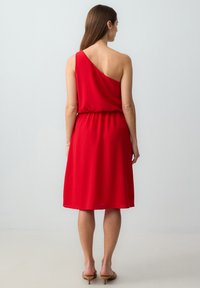 Rode, mouwloze jurk met een asymmetrische halslijn, een geraffineerde taille en een vloeiende rok die net onder de knie valt. Soepele stoftextuur.