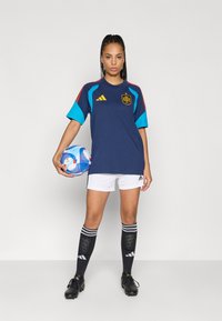 Γυναίκα με μπλε ποδοσφαιρική φανέλα της Ισπανίας, άσπρα σορτς, μαύρες κάλτσες και παπούτσια ποδοσφαίρου, κρατάει μια μπλε και άσπρη μπάλα ποδοσφαίρου, στέκεται σε απλό φόντο.