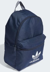 Jūronus mėlyną kuprinę su apvalia forma, priekinė zip kišenė ir balta Adidas logo. Yra tekstūruota medžiaga ir dryžuota rankena.