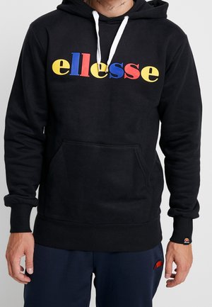 Man met een zwarte hoodie met een multicolore "ellesse"-logo, witte trekkoorden, een voorzak en een broek in marineblauw met een klein rood logo.