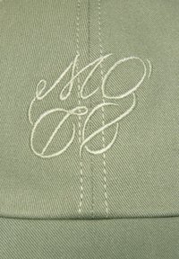 Lichtgroene pet met geborduurde initialen in een vloeiende lettertype. De stof heeft een gestructureerde, ribbelige uitstraling met zichtbare stiksels.