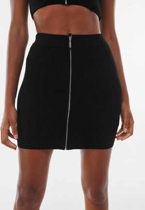 Femme portant une mini-jupe noire côtelée et un crop top assorti, tous deux avec des fermetures éclair visibles à l'avant, debout devant un fond uni.