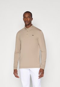 Garš sleeve polo krekls beigā audumā. Iekļauj apkakli, trīs pogas un mazu melnu logotipu uz krūšu. Vienkāršs dizains, gluda tekstūra.