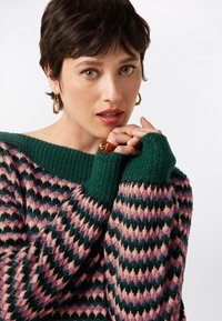 Knitted sweater with a green ribbed collar, featuring a diamond pattern in pink, purple, and cream. Fitted cuffs extend to the wrists.