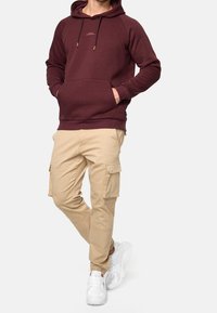Sweat à capuche bordeaux avec cordon de serrage, poche kangourou ; assorti à un pantalon cargo beige avec poches latérales ; baskets blanches.