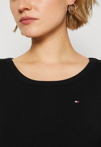 Schwarzes, geripptes Oberteil mit rundem Halsausschnitt, das ein kleines, mehrfarbiges Logo auf der linken Seite zeigt. Strukturierter Stoff mit figurbetontem Design.