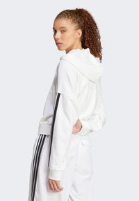 Weißer Hoodie mit einer Vorder Tasche und schwarzen Seitenstreifen. Weicher Stoff, entspannte Passform und Kapuze mit Kordelzug. Modell von hinten betrachtet.