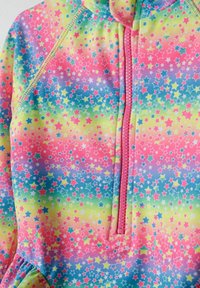 Rash guard aux couleurs vives avec un motif d'étoiles multicolores sur un fond dégradé, doté d'une fermeture éclair à l'avant et d'un tissu texturé.
