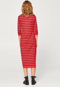 Robe longue rouge avec des rayures horizontales noires, manches 3/4, silhouette ajustée et col rond. Le tissu semble doux et texturé. Bottes noires à la cheville.