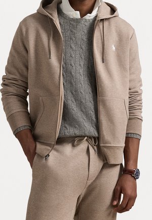Felpa beige con zip frontale e coulisse, abbinate a un maglione grigio a trecce e pantaloni beige coordinati. Design semplice, texture morbida.