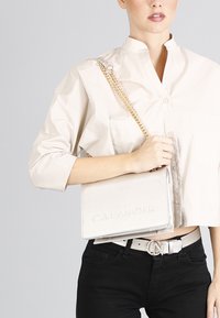 Borsa a tracolla beige con tracolla a catena, texture liscia, logo impresso "CAFÉ NOIR" e forma rettangolare elegante.