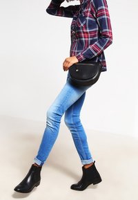 Borsa a tracolla in pelle nera con forma curva; abbinata a una camicia a quadri in blu navy, rosso e bianco, e a jeans skinny azzurri.