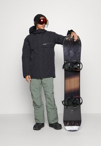 Schwarze Schneejacke mit zwei Fronttaschen, grüne Schneehose und schwarze Schneestiefel; hält ein gemustertes Snowboard mit schwarzen Bindungen.
