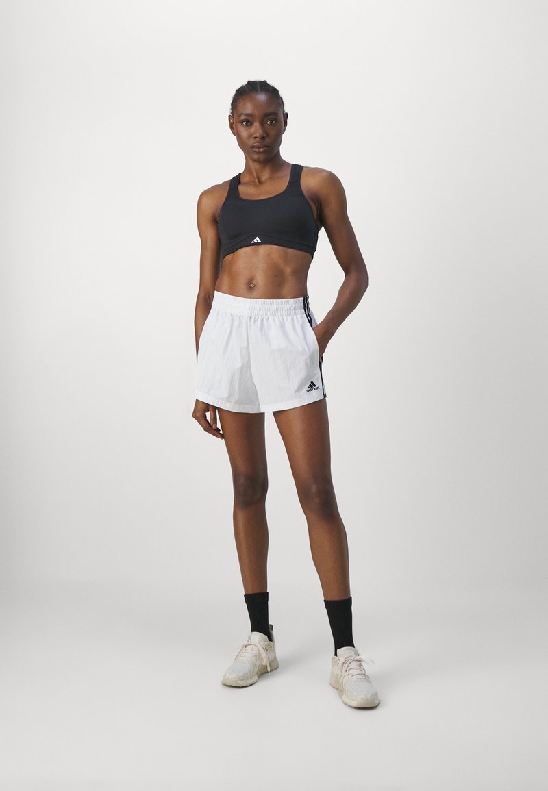 Reggiseno sportivo nero e pantaloni corti bianchi con vita elasticizzata e dettagli neri. Modella in calzini neri e sneakers chiare, in piedi.
