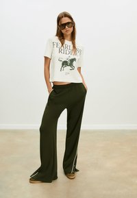 Hvid crop T-shirt med "FEARLESS RIDER" grafik i sort, parret med løstsiddende mørkegrønne bukser med hvide sidelænger.