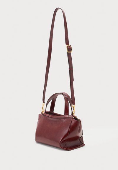Sac à main en cuir rouge foncé avec des poignées courtes et une longue bandoulière réglable avec boucles dorées sur un fond uni.