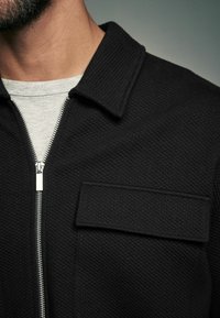 Sort jakke med lynlås lavet af tekstureret stof, en slå-om-krave og en påsyet lomme. Sølvfarvet lynlåshardware synlig foran.