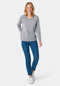 Graues Langarmshirt aus weichem Stoff, runder Halsausschnitt, kombiniert mit blauen Jeans und weißen Sneakern. Stehende Pose mit den Händen in den Hüften.