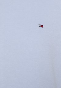 Camiseta de algodón azul claro con un pequeño logo bordado en rojo, blanco y negro, que muestra una textura suave y un cuello redondo.