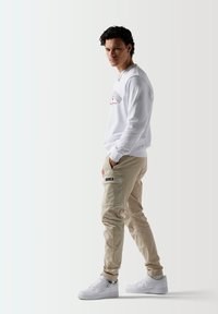 Sudadera blanca con un gráfico rojo, pantalones cargo beige con bolsillos laterales y zapatillas blancas. El fondo neutro realza el enfoque en el conjunto.