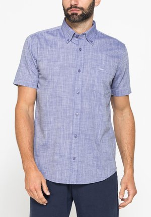 Uomo che indossa una camicia azzurro chiaro a maniche corte con taschino anteriore e pantaloni blu scuro su sfondo bianco.