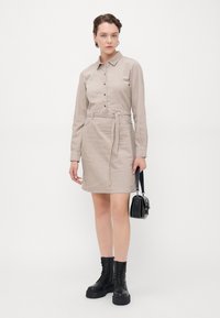 Abito camicia beige realizzato in tessuto testurizzato, caratterizzato da un colletto, chiusura con bottoni, vita con cintura e due tasche frontali. Abbinato a stivali neri e una borsa.